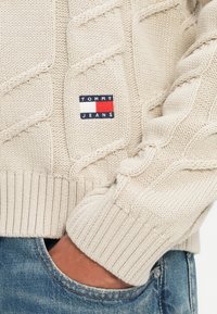 Béžový sveter z pleteniny s rebrovanými manžetami, s malou obdĺžnikovou nášivkou loga "TOMMY JEANS" v námorníckej, bielej a červenej farbe na hrudi.