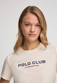Krämfärgad bomulls-t-shirt med korta ärmar, med svart och röd broderad text "POLO CLUB ESTABLISHED 2012" framtill.