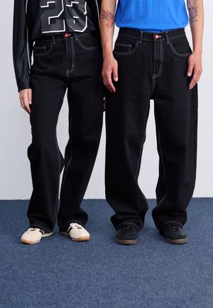 Jeans neri larghi con dettagli in cucitura bianca, indossati con camicie a contrasto e calzature casual. I jeans presentano un'altezza media in vita e un design a cinque tasche.