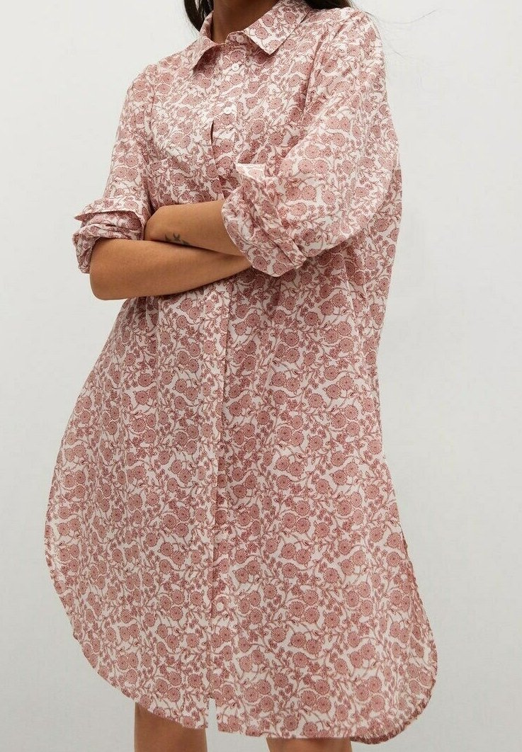 Femme portant une robe-chemise ample à imprimé floral rose et blanc, manches retroussées et col, debout, bras croisés.