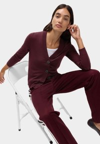 Bordowy kardigan z dekoltem w serek, trzema guzikami i ściągaczami, w zestawieniu z pasującymi bordowymi spodniami z białymi bocznymi paskami.