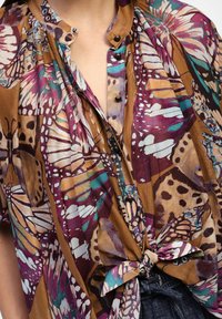 Blusa multicolore con stampa di farfalla in viola, teal e marrone. Include dettagli con bottoni e una parte frontale annodata per una vestibilità comoda.