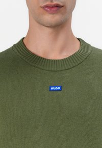 Green knitted sweater with a ribbed neckline and a blue rectangular logo labeled "HUGO" on the chest, showcasing smooth texture.