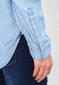 Ljusblå chambrayskjorta med knäppte ärmar, med rak passform och en slät textur, tillsammans med mörka denimjeans.