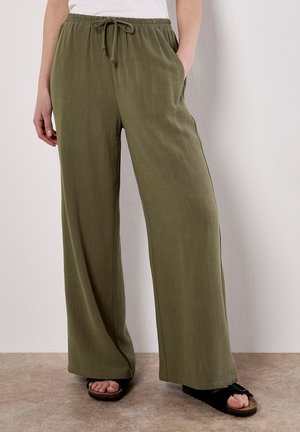 Olivgrüne weite Hosen aus weichem Stoff, mit einem Zugbandbund, Seitentaschen und einer glatten Textur. Kombiniert mit schwarzen Sandalen.