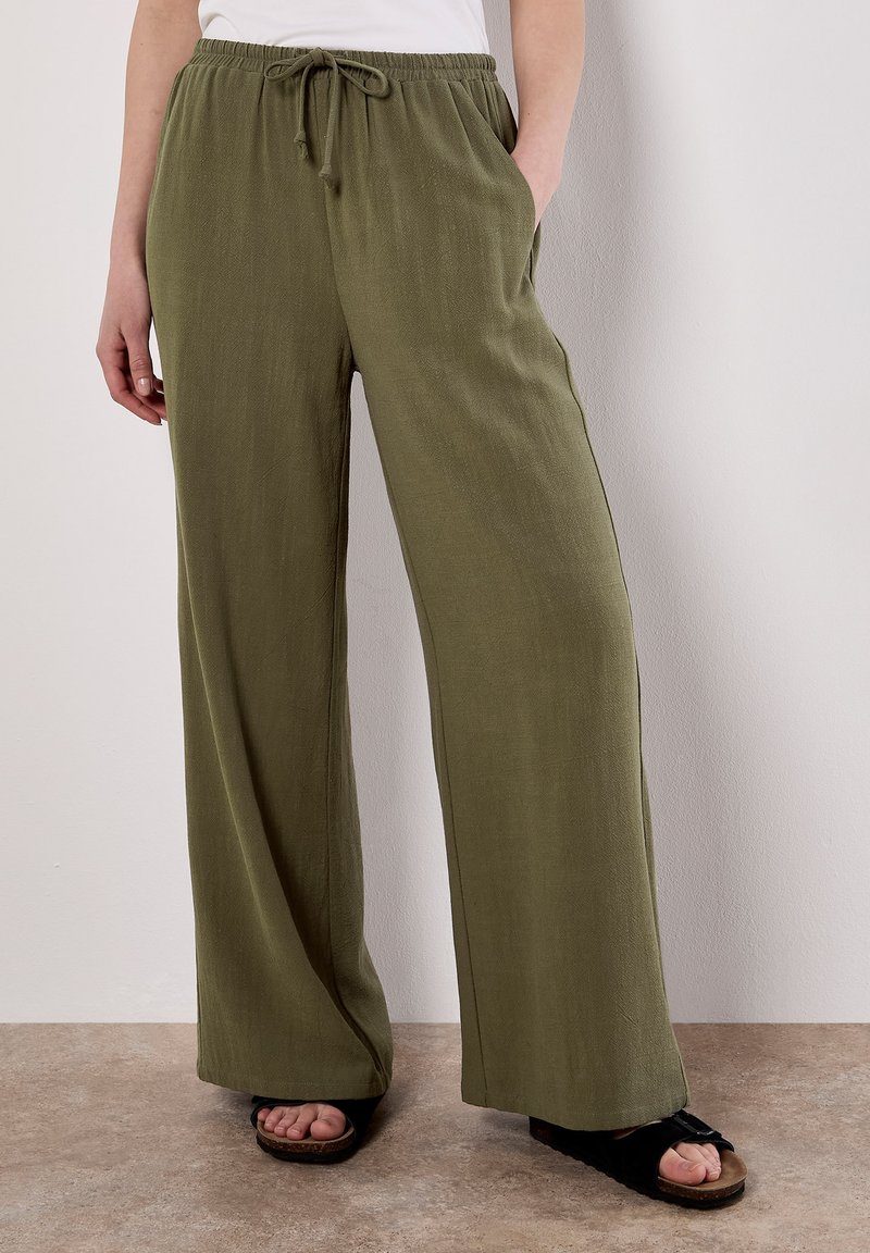 Pantaloni a gamba larga verde oliva realizzati in un tessuto morbido, con vita regolabile, tasche laterali e una superficie liscia. Abbinati a sandali neri.