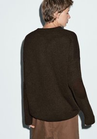 Maglione lavorato a maglia marrone, con scollatura rotonda, maniche lunghe e toppe sui gomiti. Il tessuto appare morbido con una vestibilità classica e bordi a coste.