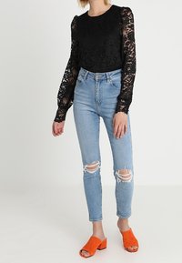 Top lungo in pizzo nero a maniche lunghe, aderente in vita; abbinato a jeans skinny strappati blu chiaro e mule aperti arancioni.
