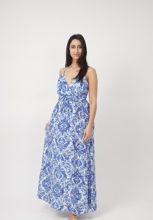 Donna in piedi che indossa un maxi vestito senza maniche con stampa floreale blu e bianca e spalline sottili, su uno sfondo chiaro e uniforme.