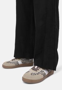 Beżowe trampki w panterkę, z białymi akcentami i brązowym sznurowadłem. Okrągły czubek i teksturowana gumowa podeszwa. Czarne, szerokie spodnie zasłaniają kostki.