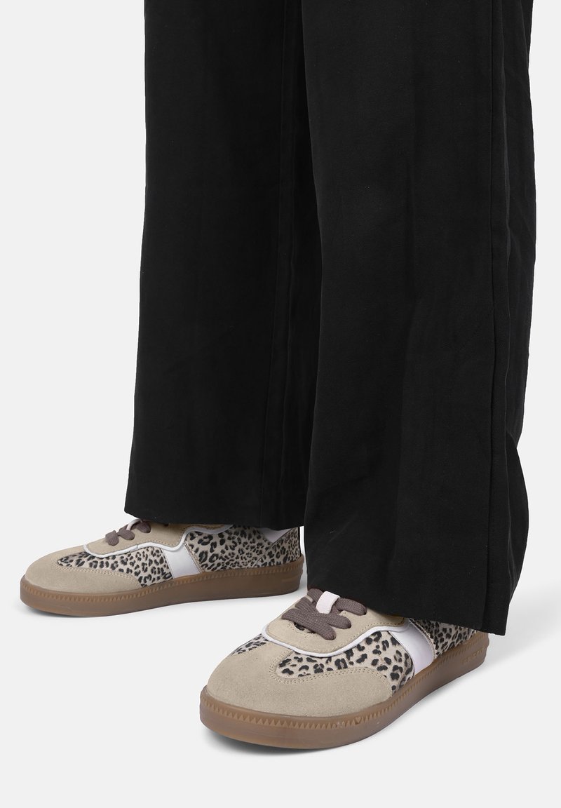 Beżowe trampki w panterkę, z białymi akcentami i brązowym sznurowadłem. Okrągły czubek i teksturowana gumowa podeszwa. Czarne, szerokie spodnie zasłaniają kostki.