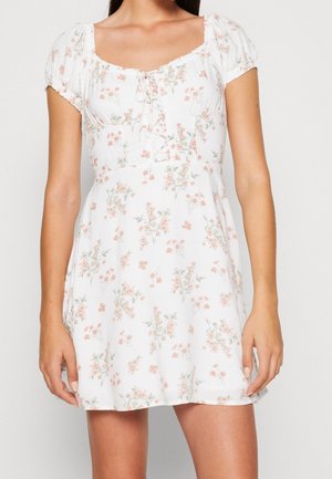 Femme portant une robe blanche courte à motif floral rose, manches bouffantes et décolleté carré avec un détail noué au niveau de la poitrine.