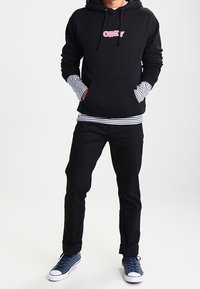 Persona con sudadera negra con el logo "OBEY", camiseta de rayas de manga larga debajo, pantalones negros y zapatillas azules, de pie con las manos en los bolsillos.