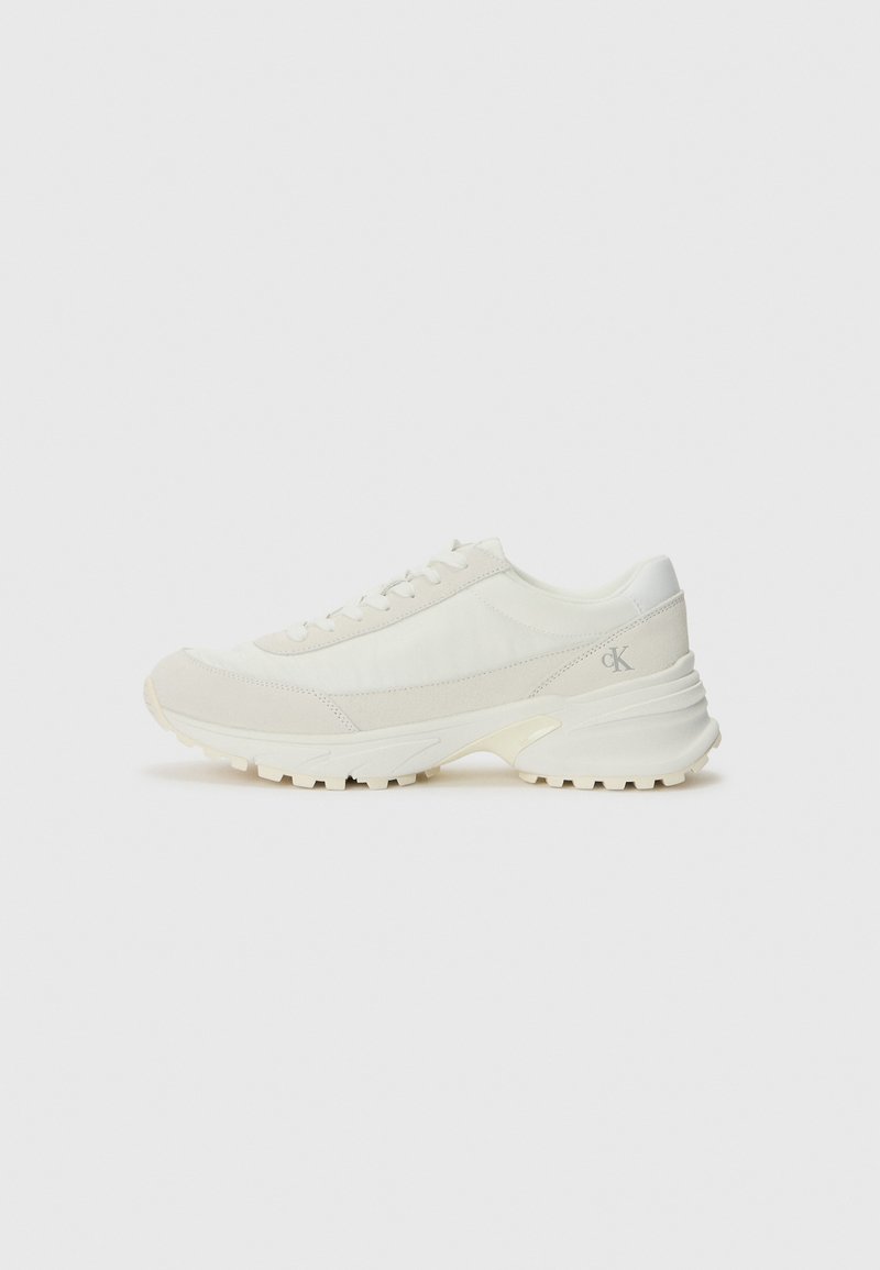 White athletic shoes with a mesh and suede upper, rounded toe, flat laces, and a textured, thick rubber sole. Logos on side and heel.