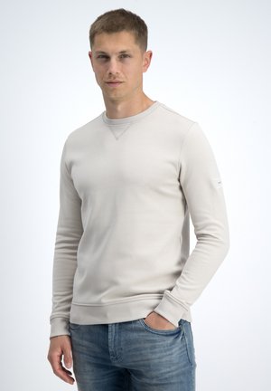 Joven de pie con una mano en el bolsillo, vistiendo sudadera beige clara y vaqueros azules, expresión facial neutral, fondo liso.