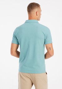 Turquoise poloshirt met korte mouwen en een kraag, gemaakt van een textuurstof. Het model draagt beige broek en toont een tattoo op de arm.