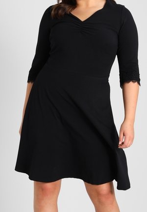 Femme portant une robe noire longueur genou avec des manches trois-quarts et une bordure en dentelle, présentant un col froncé et une jupe évasée.