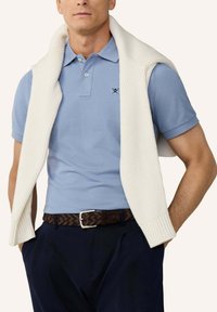 Mężczyzna w jasnoniebieskiej koszulce polo z małym logo skrzyżowanych wioseł, granatowych spodniach, brązowym plecionym pasku oraz białym dzianinowym swetrze zarzuconym na ramiona.