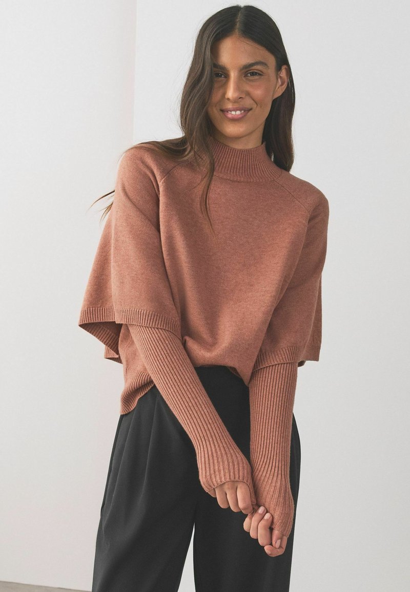 Nő mosolyog, barna bordázott garbó pulóvert visel, hosszú ujjakkal és fekete bőszárú nadrágot, fehér fal előtt áll.