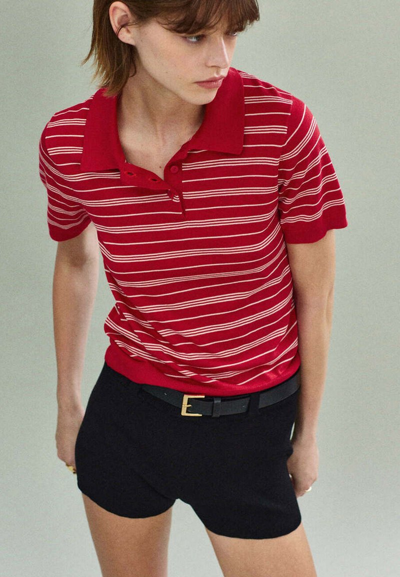 Jeune femme portant un polo rayé rouge et blanc rentré dans un short noir avec une ceinture noire, regardant vers sa gauche.
