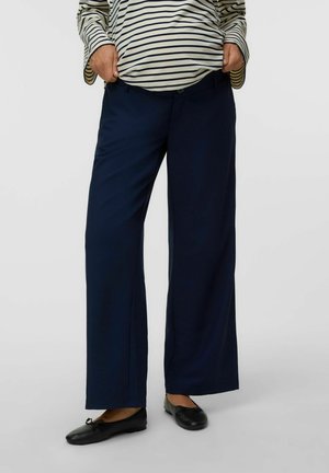 Personne portant un haut rayé crème et noir, un pantalon bleu foncé à jambes larges et des ballerines noires, les mains dans les poches, debout bien droit.