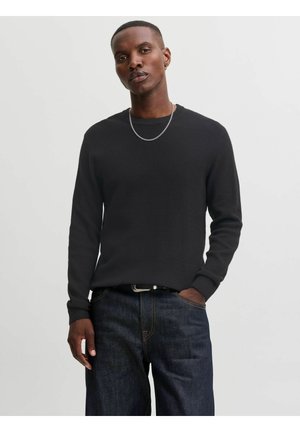 Homme en pull à manches longues noir texturé et jean bleu foncé, portant un collier chaîne fine et une main dans la poche, sur fond gris uni.