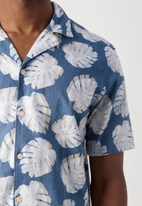 Homme portant une chemise à manches courtes bleue avec de gros imprimés de feuilles tropicales blanches et des boutons beige, visible du cou jusqu'au milieu du torse.