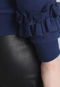 Navy rukáv s volánkovým lemem přes hladkou černou koženou tkaninu, zobrazuje zápěstí a dolní část ruky.