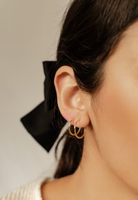 Orecchini a cerchio dorati indossati all'orecchio sinistro, con una superficie liscia e lucida e due cerchi impilati uno vicino all'altro. È visibile un nastro nero.