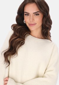 Suéter de punto crema con cuello redondo y mangas largas, ligeramente holgadas. La textura es suave y acogedora, con un patrón sutil.