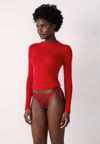 Rotes Langarmoberteil aus glattem Stoff, kombiniert mit durchsichtigen roten Spitzenunterhosen mit floralem Stickerei und seitlichen Bindedetails.