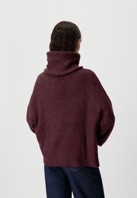 Burgundský svetr s vysokým, skládacím límcem; pletená textura; volný střih; široké rukávy; nošený přes modré džíny.
