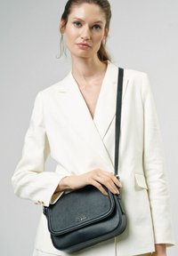 Czarna skórzana torba na ramię o teksturowanej powierzchni, z regulowanym paskiem i złotym logo, trzymana przez osobę w białym żakiecie.