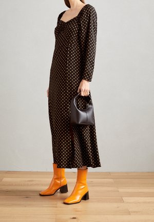 Frau trägt ein schwarzes Midikleid mit gelben Punkten, hält eine kleine schwarze Handtasche und trägt leuchtend orangefarbene Stiefeletten mit Blockabsätzen.