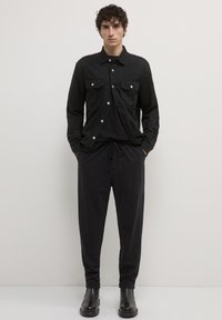 Mann, der gegen eine weiße Wand steht, trägt eine schwarze Knopfjacke, schwarze Hosen mit Kordelzug und schwarze Stiefel, die Hände in den Taschen.