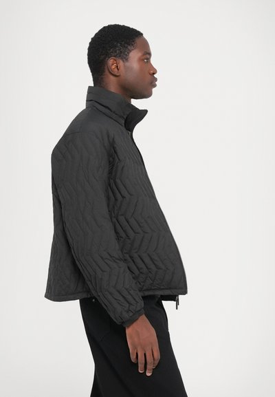 Man facing right wearing a black quilted jacket with high collar and black pants against plain light background.