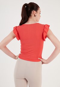 Top rosso con texture e maniche corte a volant; design aderente. Indossato con leggings beige, modello di schiena verso la fotocamera, mani sui fianchi.