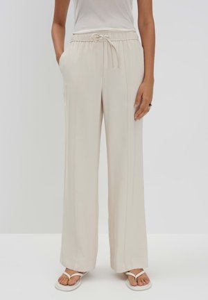 Kobieta ubrana w luźne beżowe spodnie na sznurek z bocznymi kieszeniami i białe sandały, stojąca na tle jednolitego jasnego tła.