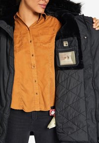 Mujer con camisa naranja de botones abre un abrigo negro acolchado de invierno con cuello forrado de piel, mostrando un bolsillo interior cosido con etiqueta.