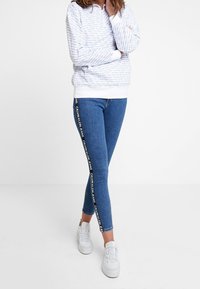 Felpa azzurro chiaro con motivo di testo, abbinata a jeans skinny blu scuro con nastro logo bianco lungo il lato, e sneakers bianche.