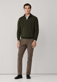 Suéter de cuello con cremallera en verde oliva con un acabado texturizado, combinado con pantalones marrones y zapatos negros. Diseño slim-fit, con hardware mínimo.