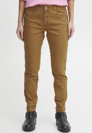 Bruna tajta jeans med femknappsstängning, bältesloopar och sidoficka med dragkedja. Tyget ser slätt ut med lite stretch. Svarta ankelstövlar synliga.