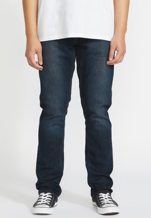 Homme portant un jean bleu foncé, un t-shirt blanc et des baskets à lacets noires et blanches, debout avec les mains relâchées le long du corps.