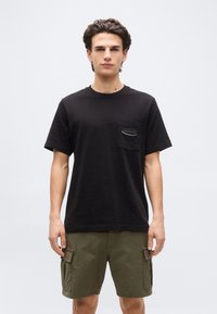 Άνδρας που στέκεται και κοιτάζει μπροστά, φοράει μαύρο T-shirt της Patagonia με τσέπη στο στήθος και χακί cargo σορτς, μπροστά σε απλό φόντο.