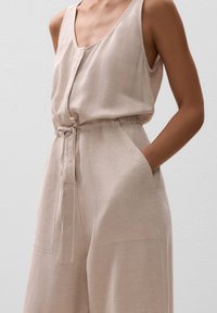 Beige Leinen-Overall mit Rundhalsausschnitt, verstellbarem Taillengurt und seitlichen Taschen, der eine lockere Passform und eine glatte Textur bietet.