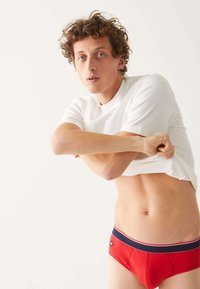 Caleçons rouges avec une ceinture bleu marine et blanche ; le mannequin porte un t-shirt blanc uni, le soulevant. Fond neutre, mise au point sur les détails du produit.