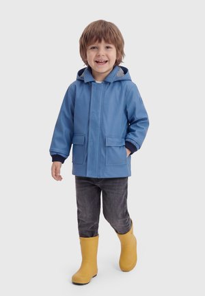 Enfant souriant portant un imperméable bleu, un pantalon foncé et des bottes de pluie jaunes marchant vers l'avant sur un fond uni.