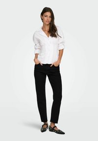 Camisa blanca de botones con hombros estructurados y textura acanalada, combinada con jeans negros de pierna recta y zapatos planos negros con tachuelas.