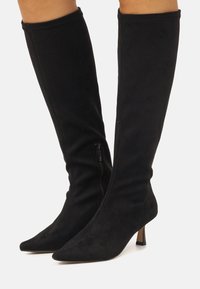 Botas negras por encima de la rodilla hechas de material de ante suave, con punta afilada, tacón delgado y un cierre lateral para facilitar su uso.