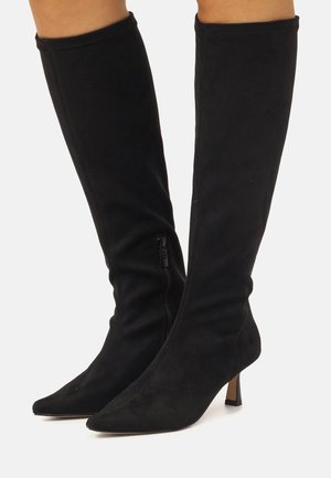 Botas negras por encima de la rodilla hechas de material de ante suave, con punta afilada, tacón delgado y un cierre lateral para facilitar su uso.